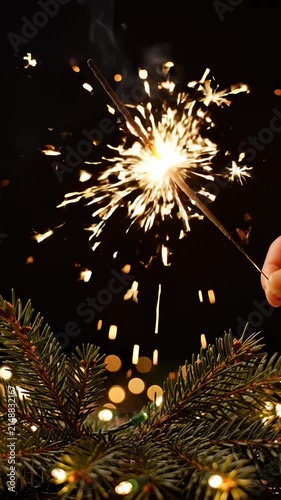 Hand Holding Burning Sparkler Above Christmas Tree Branches With Bokeh Lights