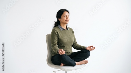 Mature Indian woman meditating in lotus position on office chair with eyes closed and serene expression