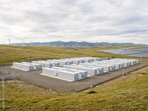 A massive battery storage facility, featuring rows of rectangular units and complemented by distant wind turbines and solar panels.