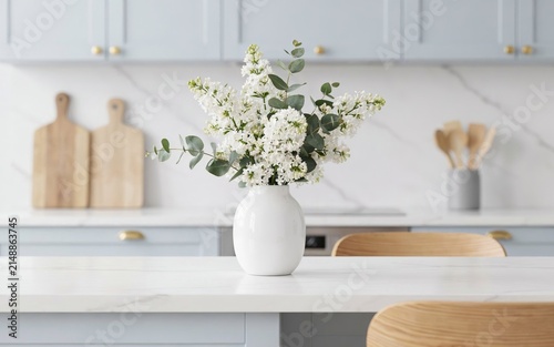 A white ceramic vase filled with a fresh bouquet of white lilac flowers and green eucalyptus leaves, placed on a white marble countertop in a bright, modern kitchen with light blue cabinets