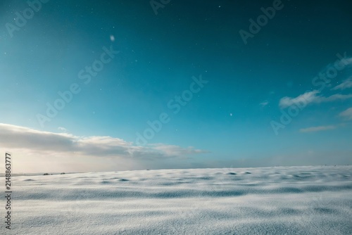 A wide, snow-covered field stretching towards a clear blue sky with a few scattered clouds and faint stars, capturing the cold and quiet atmosphere of a winter landscape during the twilight hour