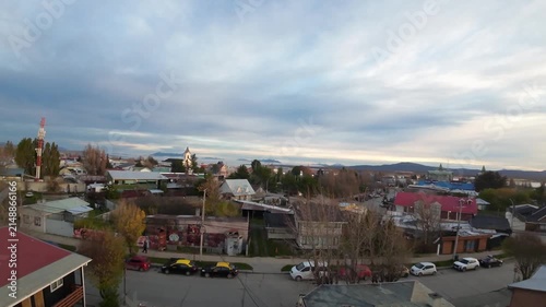 FPV drone footage flying low over colorful rooftops toward the Parroquia María Auxiliadora and Plaza Arturo Prat, with the fjord and mountains beyond Puerto Natales, Chile