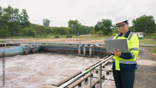 Asian woman worker checking industrial water treatment facility and wastewater basin with laptop and clipboard. Environmental protection, sanitation and engineering concept.