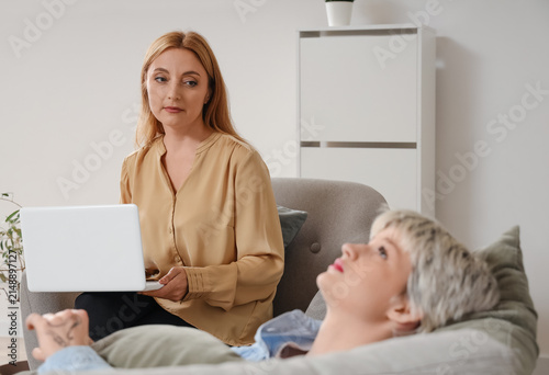 Female psychologist with laptop and sad teenage girl lying on sofa in office