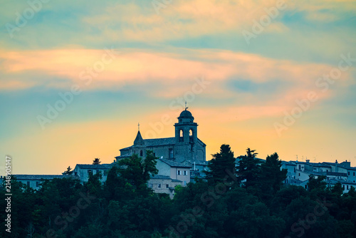 Historic church of San Michele Arcangelo in Mondaino illuminated by soft sunset light above forested hills on the Marche Romagna border showing peaceful Italian landscape