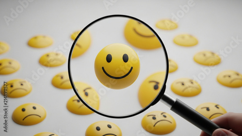 Magnifying glass held by hand focusing on a single yellow 3D smiling emoji face among many blurred frowning and sad icons on a light gray surface, concept of finding positivity.