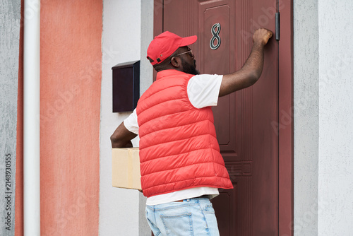 Foto African American young adult courier knocking on door while holding cardboard box, delivering package to customer home