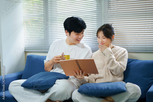Konstfotografi Young happy couple sits together on a cozy blue couch in their living room, looking at a memory book together and smiling while reminiscing about cherished times and drinking coffee