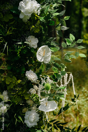 The side of a festive arch made of live plants and white lilies is placed next to a white table outdoors in a dense forest