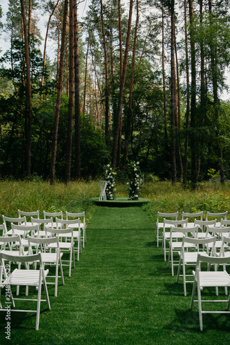 The wedding ceremony venue is located in a dense forest on a well-groomed lawn with white chairs for guests and a wedding arch made of live plants and flowers
