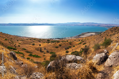 Konstfotografi Wide panoramic landscape of Lake Kinneret (Sea of Galilee) viewed from the Golan Heights, featuring dry golden hills, rocky foreground, blue water, and a clear summer sky