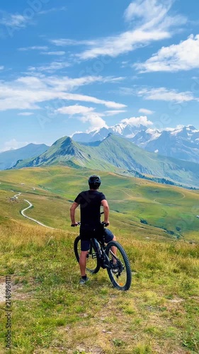 Male cyclist  with mountain electric bike on grassy hill looking at majestic alpine peaks under blue sky