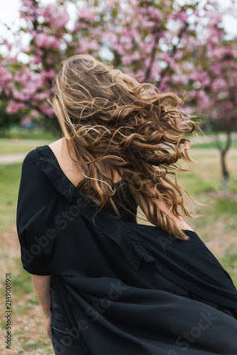 Back view of woman with long curly hair waving in the wind against blooming sakura background.