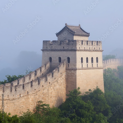 A majestic segment of the Great Wall is shrouded in morning mist, surrounded by lush greenery, This serene image is ideal for travel, history, or cultural themes in various media,