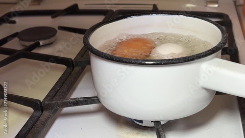 Close-up boiling eggs in enameled ladle on gas stove, brown and white chicken eggs simmering in bubbling water, steam rising, white