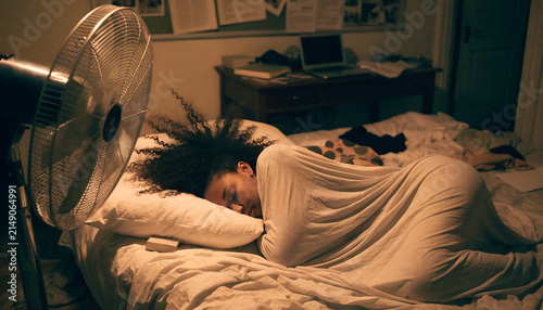 Young Black woman sleeping peacefully in a comfortable bed during a hot summer night, refreshed by the gentle breeze of a modern pedestal fan in a cozy dark bedroom.