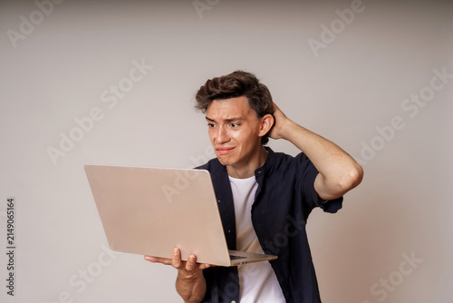 Confused young man looking at laptop screen while scratching his head on neutral background