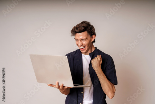 Young man celebrating success while using laptop against neutral background