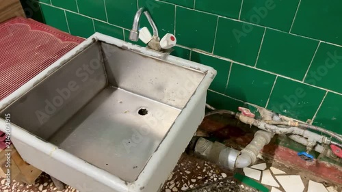 Rusty iron foot sink under faucet, set against green ceramic tiles, exposed pipes and valve, stained basin floor with clogged