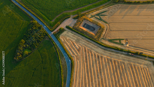 Aerial view of golden wheat fields during harvest season with a combine harvester working near a road and a small pond in Enderby, England, United Kingdom.