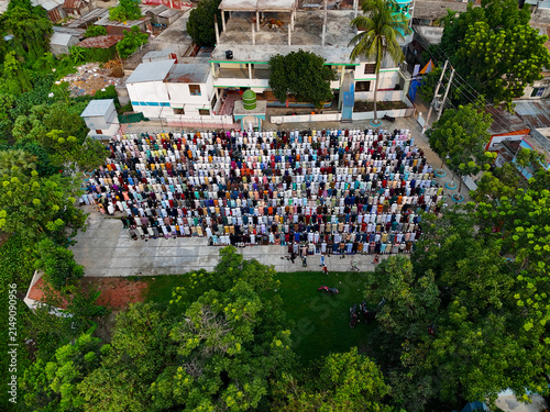 Aerial view of a large crowd of men gathered for outdoor prayer in an open courtyard near a mosque with a green dome in Kushtia, Khulna Division, Bangladesh.