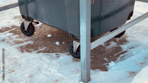 Plastic trash can behind fence, snow. Blue gray plastic dumpster chained to metal rail beside brick wall, snowbanks piled