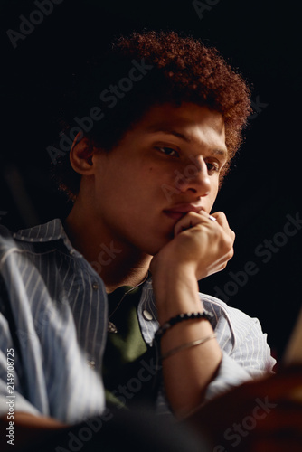 Canvas Print Young adult male student sitting in dark theater, holding chin in hand while reading script