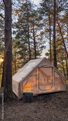 Glamping tent glowing at sunset among tall pine trees inside quiet forest campsite, warm interior light, soft sky behind branches, rustic outdoor retreat in nature