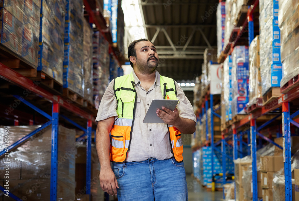 Obraz premium Warehouse worker using a tablet while standing in a large distribution center aisle stacked with boxes