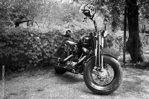 Black and white photograph of a classic  motorcycle parked on a gravel path in a quiet garden surrounded by lush vegetation. Two vintage-style helmets rest on the motorcycle, emphasizin