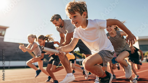 Group of children running a relay race on an athletics track, passing baton with a coach supervising them