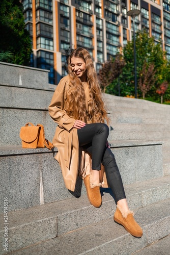 Young smiling woman in tan trench coat seated on stone steps in urban cityscape with beige handbag and modern buildings behind, stylish outdoor portrait, with woman, female, adult, smile detail