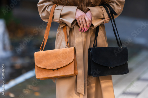 Beige trench coat styled on woman holding orange and black handbags with urban fashion pose and visible accessory details for street style concept, with handbag, fashion, woman, coat detail