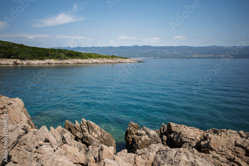 Foto Sharp limestone rocks in the foreground along the pristine coastline of Krk Island, Croatia, overlooking clear turquoise Adriatic waters, a green pine-covered peninsula under a bright blue sky