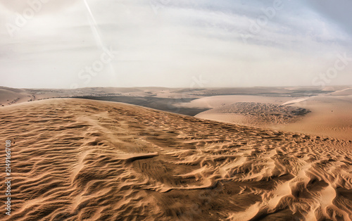Canvas Print Expansive desert sand dunes with intricate patterns shaped by wind on the foreground dunes and distant rolling hills under a hazy, bright sky