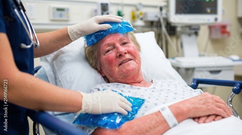 Elderly female patient in hospital bed receiving treatment for heatstroke. Nurse applying blue ice packs to flushed senior woman's forehead and chest. Medical care and fever reduction