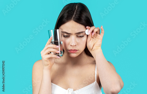 Woman take some pills, holds glass of water, isolated on blue. Young woman taking pill against headache. Female taking a pill with a glass of water. Woman taking drugs to relieve headache