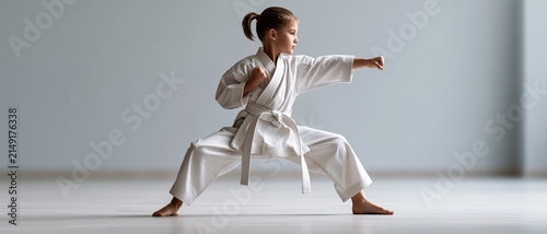 A young girl in white gi practices a strong karate punch stance in an empty dojo with a neutral background and wooden floor