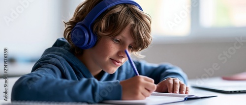 A young boy wearing blue headphones and a blue hoodie is sitting at a desk writing attentively in a notebook with soft natural light coming through a window