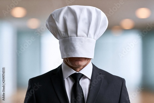 Man in formal black suit and tie wearing a chef's hat covering his eyes in an indoor blurred background environment