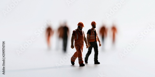 Two miniature construction workers walking forward in safety gear with blurred colleagues in the background on a white surface