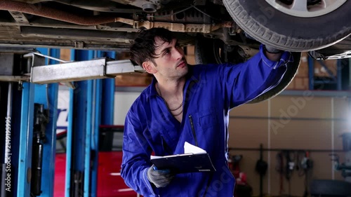Focused mechanic checking vehicle tire with happy expression repair shop, working automotive maintenance status digital tablet, professional