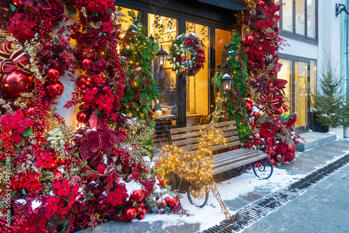 Festive Christmas decoration of a restaurant facade in Moscow with red ornaments, baubles, poinsettia flowers, evergreen garlands, illuminated reindeer sculpture, a wooden bench, and winter snow.