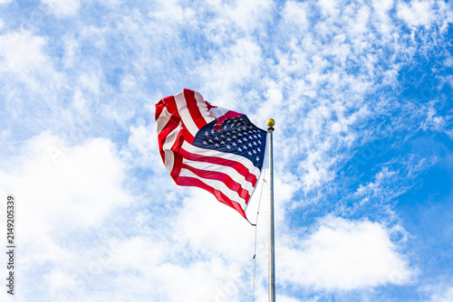 United States national flag waving on a metal flagpole against a bright blue sky with textured white clouds in broad daylight