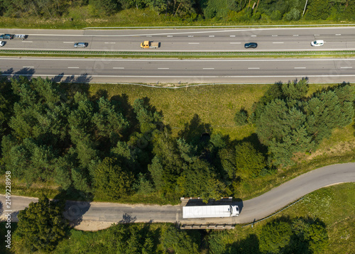 Aerial view of a highway with moving cars and a white truck on a secondary road surrounded by dense green forest in Vilnius, Vilnius County, Lithuania.