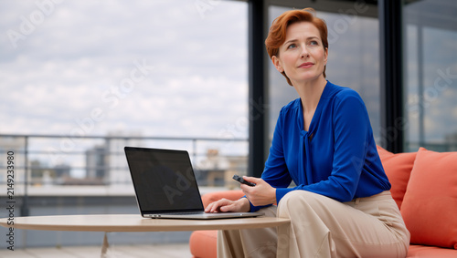 Thoughtful mature businesswoman with laptop on outdoor balcony. Redhead female executive holding smartphone and looking away on city terrace. Remote work and corporate lifestyle concept