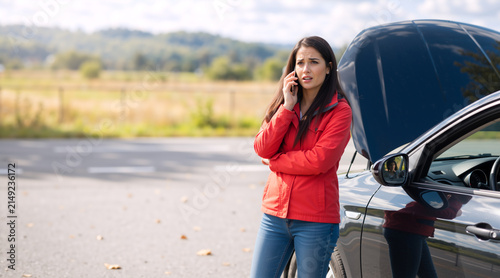 Worried young woman calling for roadside assistance next to a broken down car. Female driver talking on the phone by a vehicle with an open hood