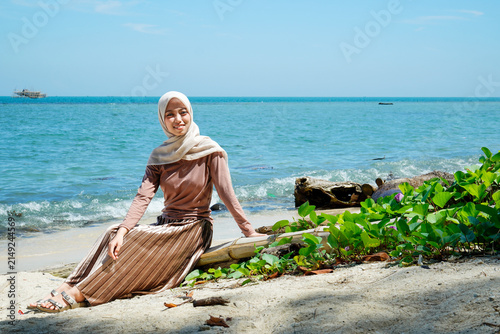 ​Wide shot of a smiling young Asian Muslim woman wearing a beige hijab, long-sleeved brown top, and pleated skirt, sitting on a bamboo log at the sandy shoreline.