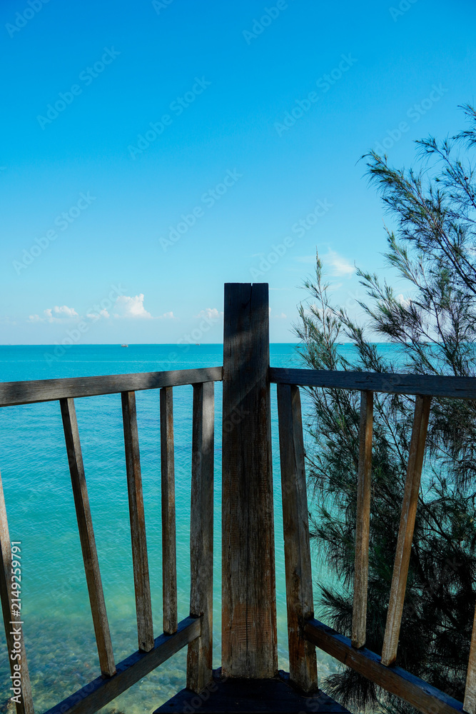 Fototapeta premium Scenic view of a rustic wooden balcony fence overlooking clear turquoise ocean water and green coastal pine trees under a bright blue sky. 