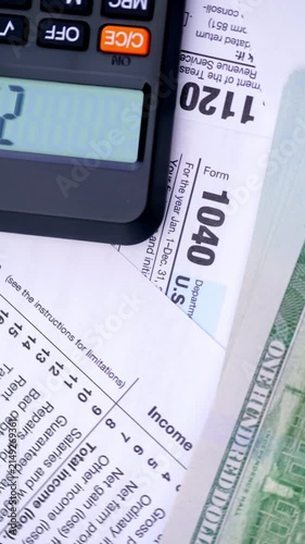 Calculating Income Tax Return. A hand enters figures on a calculator beside tax forms, dollar bills, and a pen.	
